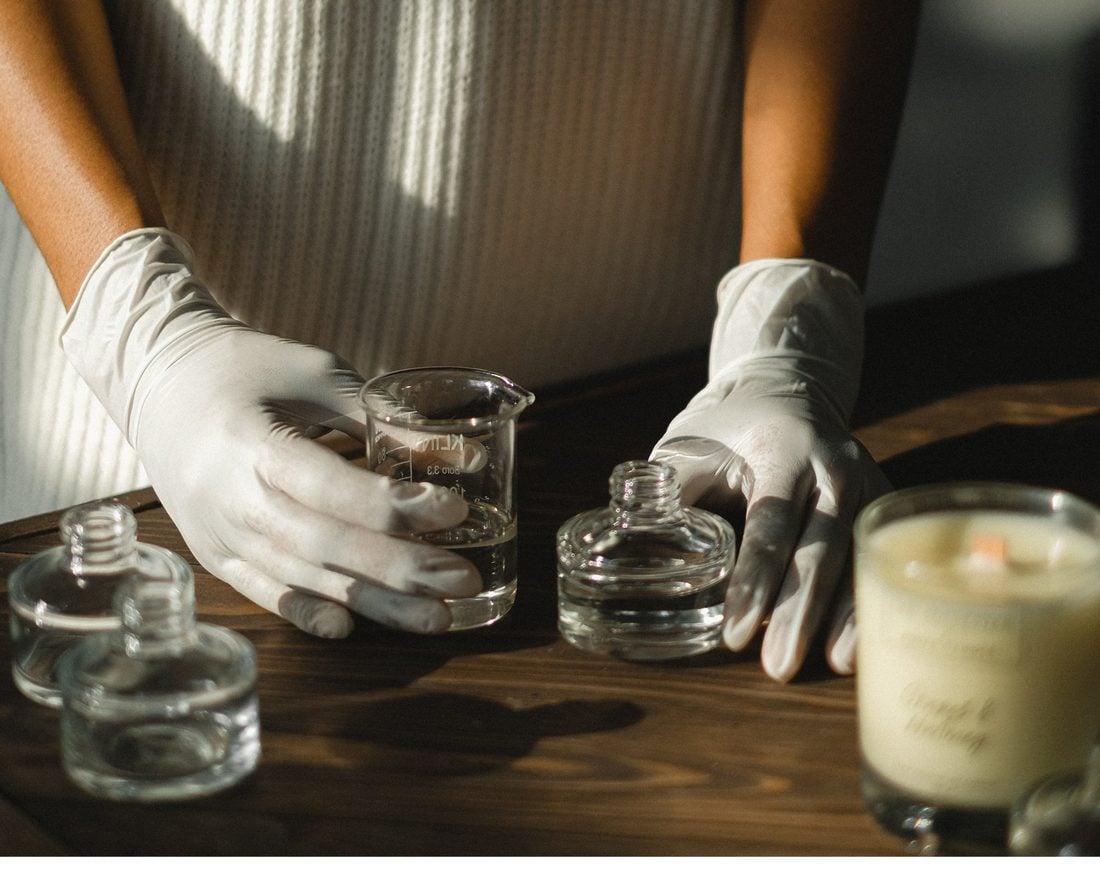 Fleur&Fume atölyesinde koku kompozisyonu — eldivenli eller, ölçü kabı ve cam parfüm şişeleri